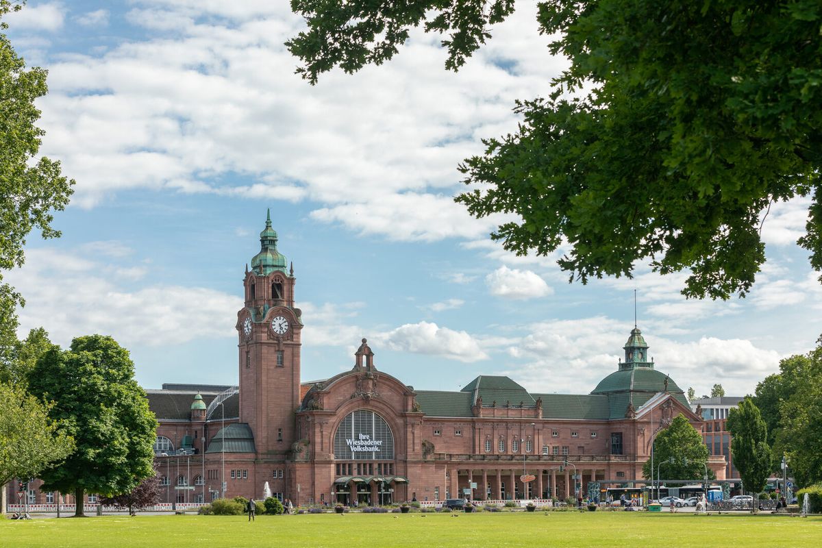 Der Wiesbadener Hauptbahnhof aus roten Sandsteinquadern mit Uhrturm, davor eine Rasenfläche