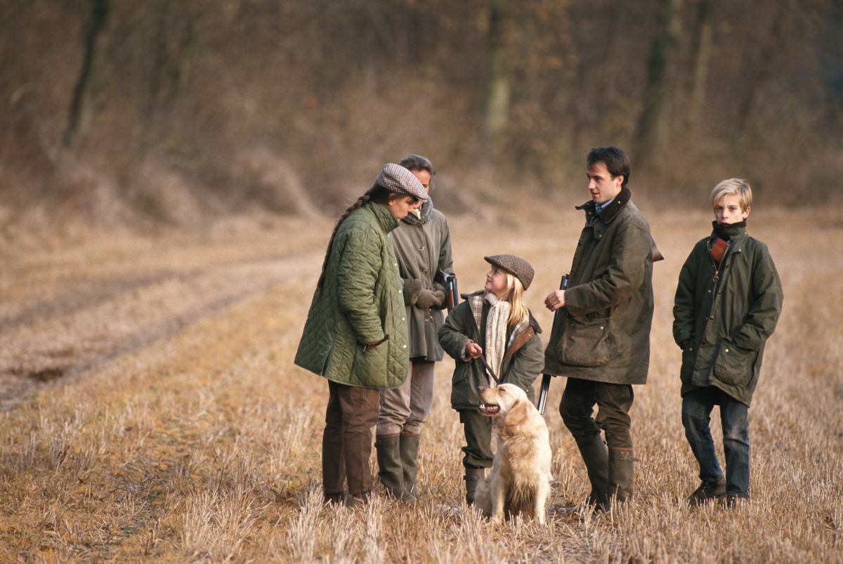 Mehrere Personen in wetterfester Jagdkleidung stehen mit einem hellen Retriever auf einer trockenen Wiese zusammen.