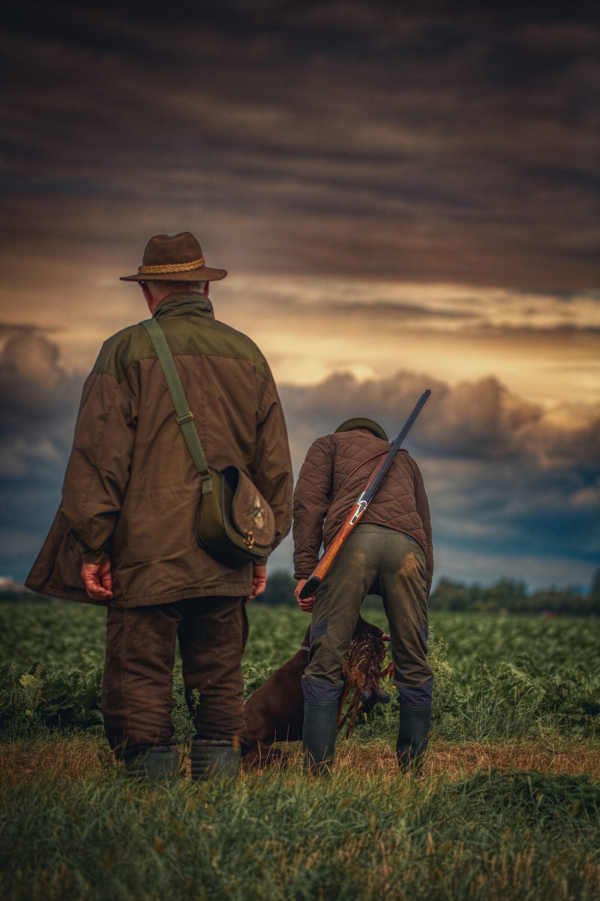 Jagdhund übergibt einem Hundeführer im Feld ein Stück Federwild, ein zweiter Jäger steht daneben