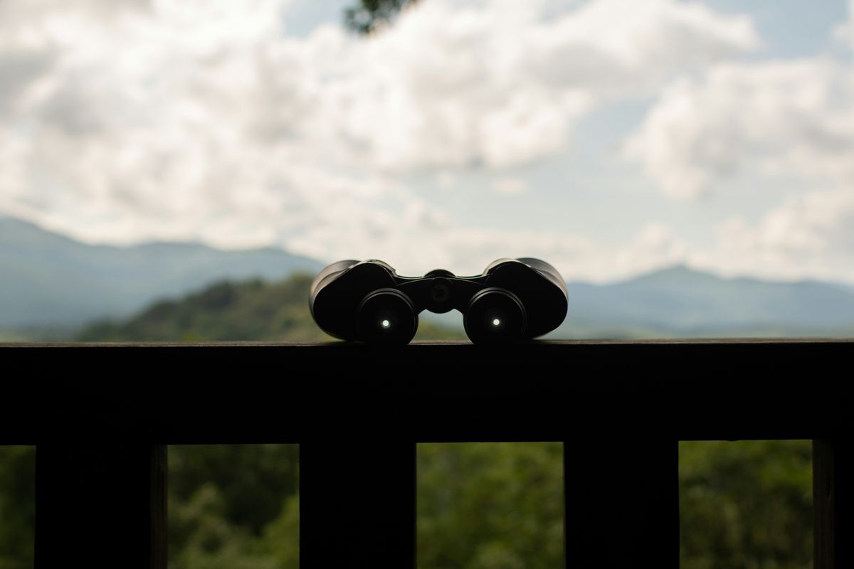 Ein schwarzes Fernglas steht silhouettenhaft auf einem Holzgeländer vor einer bewölkten Berglandschaft.